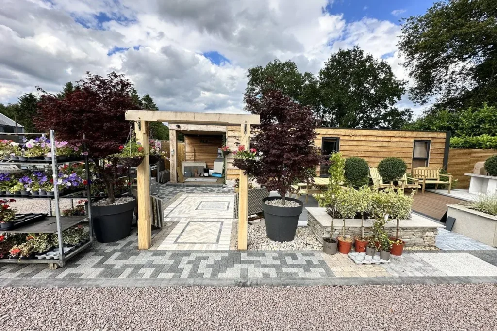 Show garden with an oak pergola framing a patterned block-paved path to a timber-clad cabin, with acers and bay trees