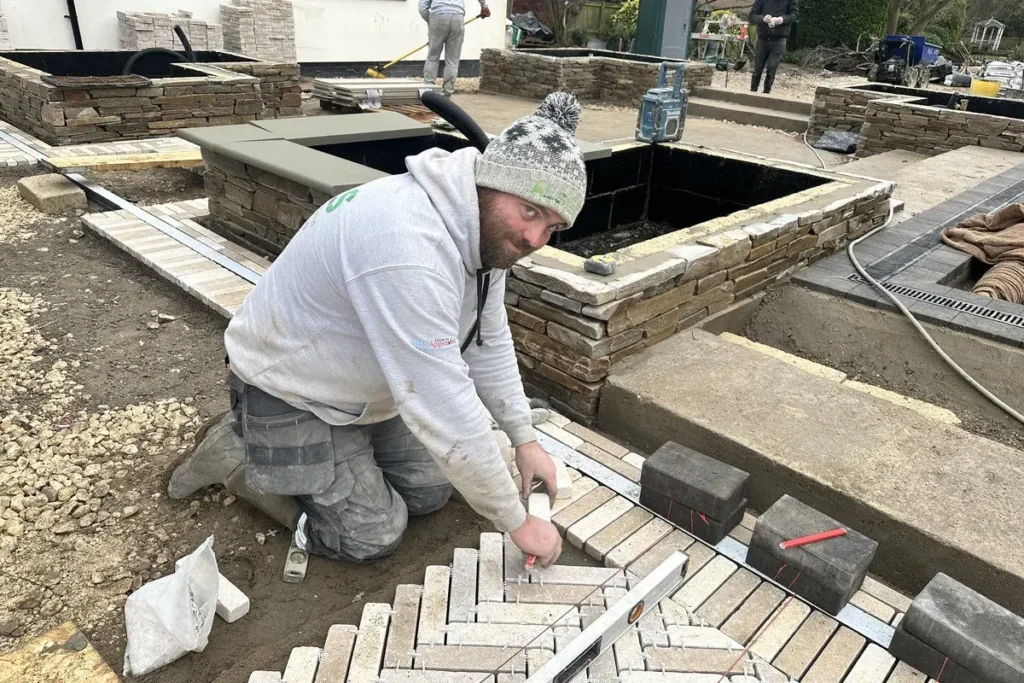 Craig Holloway cutting and laying buff herringbone pavers beside dry-stone raised beds