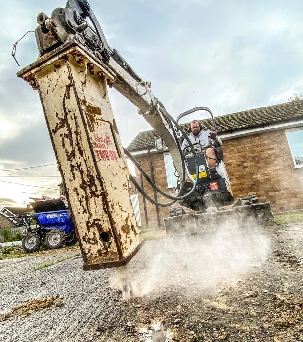 Mini digger with a hydraulic breaker breaking out an old driveway, operator in the cab