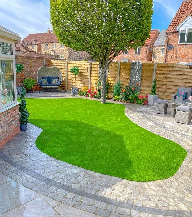 Kidney-shaped artificial grass lawn with a sett border, a round-headed tree and seating along a timber fence