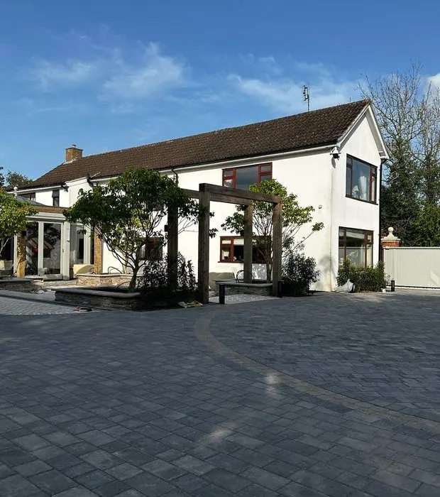Large grey tumbled block-paved driveway with a curved sett band in front of a white house with oak pergolas