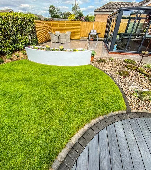 Lawn seen from a curved composite deck, with a white curved planter, patio dining area and glass garden room