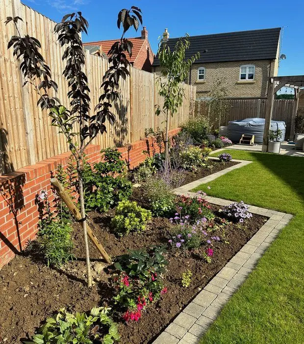 Planted border with young trees and perennials edged in buff blocks against a fence on a red-brick plinth