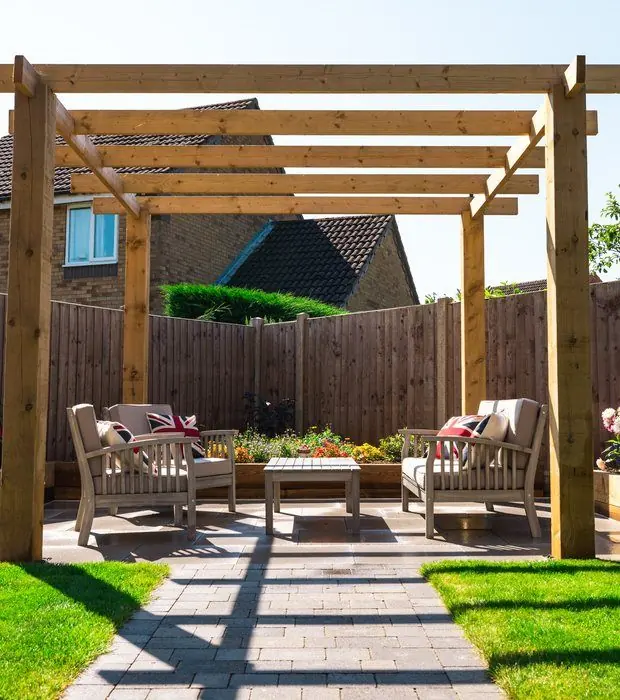 Timber pergola framing two wooden armchairs on grey paving between raised sleeper beds of bedding plants