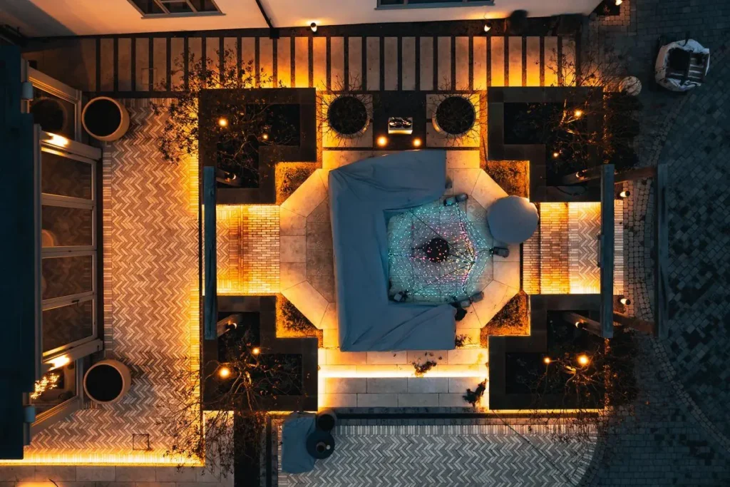Overhead dusk view of a cross-shaped terrace with lit raised beds and herringbone paths