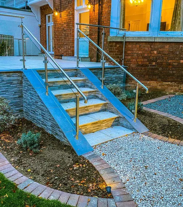 Porcelain steps with a glass balustrade up to a raised terrace at dusk, with lit planting