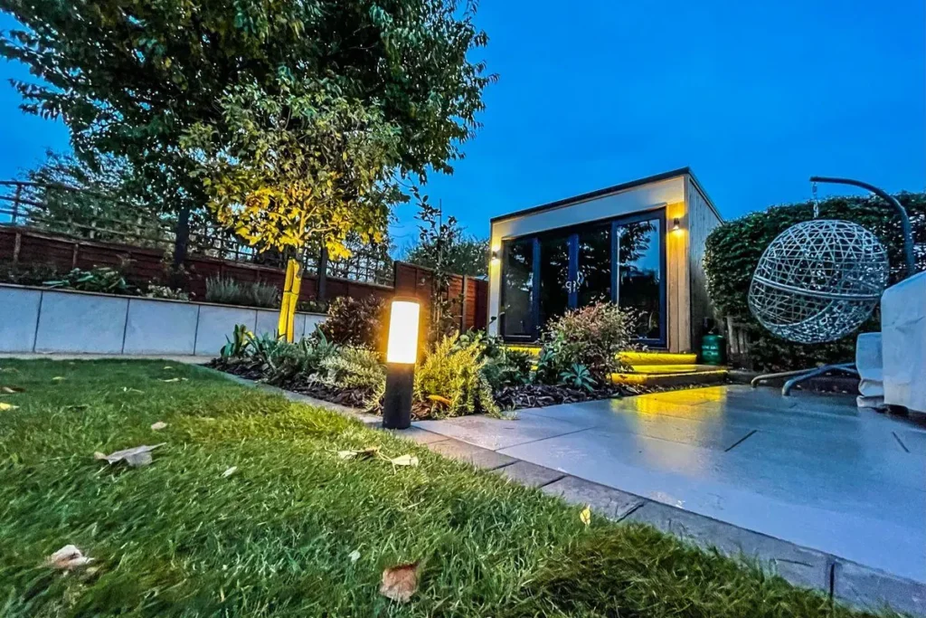 Modern garden room with wall lights, a glowing bollard and an uplit tree at dusk