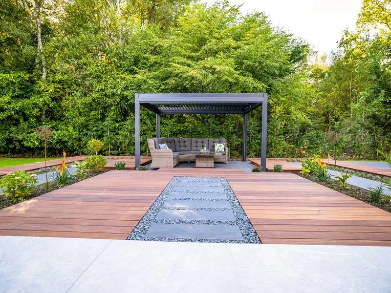 Hardwood decking with a central strip of grey stepping slabs in pebbles leading to a dark louvred pergola