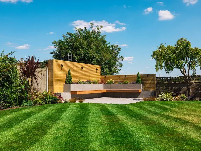 Striped lawn leading to a porcelain patio with an L-shaped floating bench, raised planters and slatted timber screen