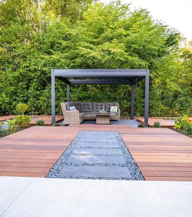 Hardwood decking with stepping slabs in pebbles leading to a dark louvred pergola with a rattan sofa