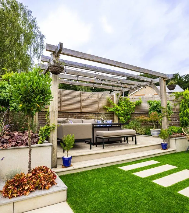 Oak pergola over a sofa platform with stepping stones across an artificial lawn and a raised bed